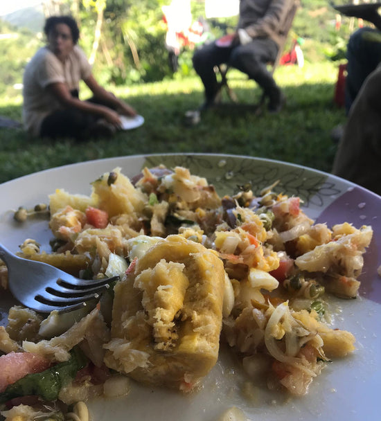 un plato de comida (habichuelas, viandas, vegetales), con otras personas en el fondo comiendo en el campo