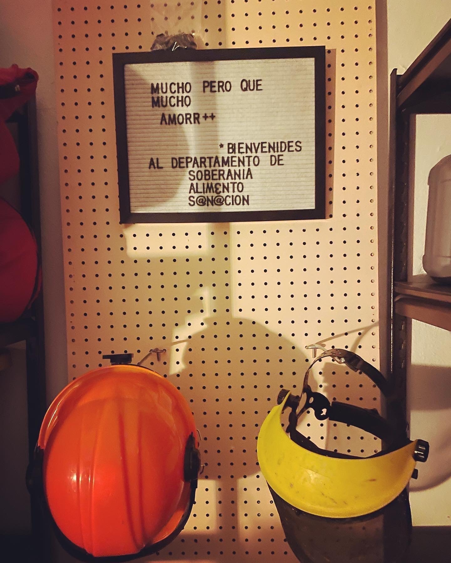 un pegboard que tiene un letrero que dice "Much pero que mucho amorr++. Bienvenides al Departamento de Soberania, Aliemnto, Sanacion". Abajo, son 2 cascos.